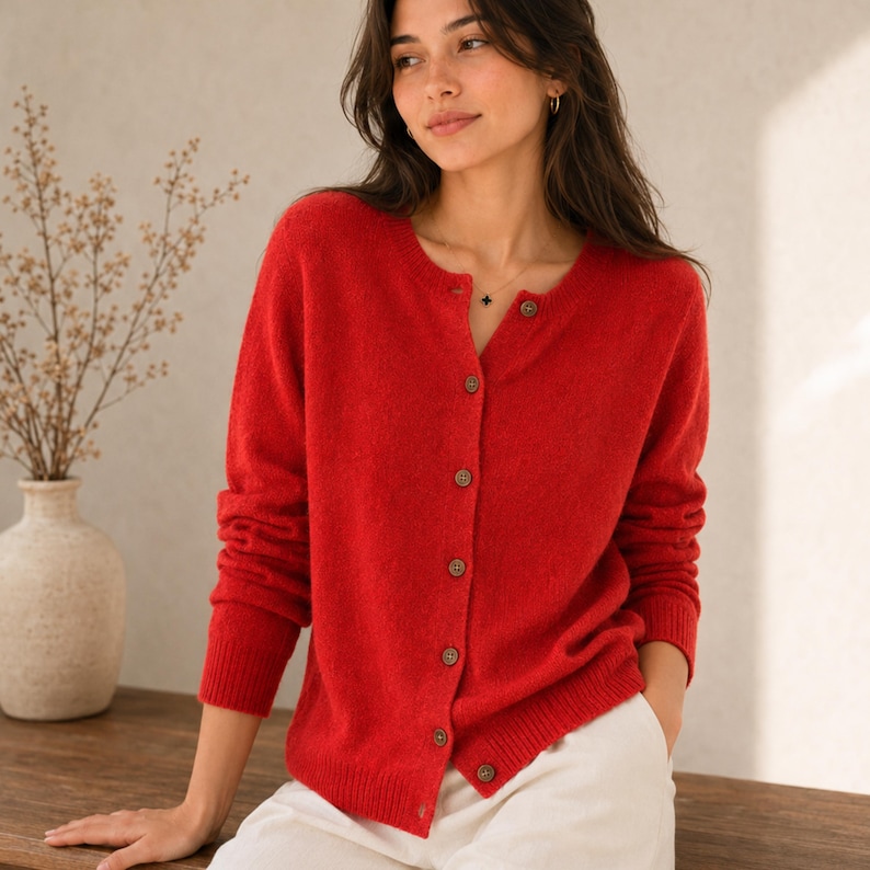 May include: A vibrant red long-sleeved knit cardigan featuring a round neckline and a vertical row of small, round, dark-coloured buttons down the front. The garment has ribbed cuffs and a ribbed hem, providing a structured fit with a soft texture.