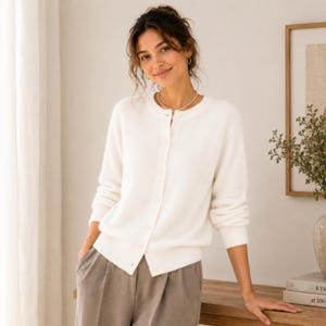 May include: A long-sleeved, off-white button-down cardigan featuring a round neckline and ribbed cuffs. The garment is paired with grey trousers. The cardigan has a soft, knit texture and is fastened with a vertical row of matching white buttons.