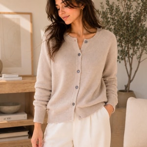 May include: A beige long-sleeved cardigan with a round neckline and a front button closure featuring five visible buttons. The knit garment has ribbed cuffs and a ribbed hem, providing a structured fit with a soft, textured appearance.