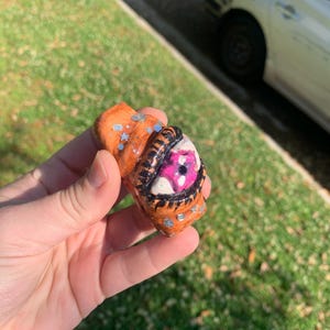May include: A handmade, orange-colored, abstract sculpture with a detailed eye design. The eye features a white iris, a pink pupil, and black outlining. Silver accents are scattered across the surface. The sculpture is held in a hand.