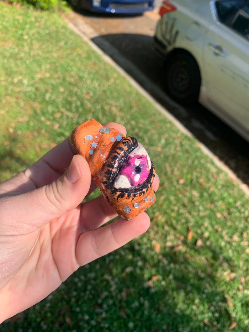 May include: A handmade, orange-colored, abstract sculpture with a detailed eye design. The eye features a white iris, a pink pupil, and black outlining. Silver accents are scattered across the surface. The sculpture is held in a hand.