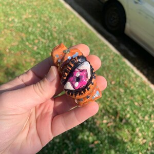 May include: A small, orange, handmade sculpture with a detailed eye design. The eye is pink and white with black outlining. The sculpture has silver glitter and star accents. The object is held in a hand.