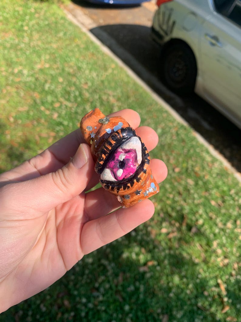 May include: A small, orange, handmade sculpture with a detailed eye design. The eye is pink and white with black outlining. The sculpture has silver glitter and star accents. The object is held in a hand.