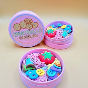 May include: Pink round boxes with lids, one labeled "Shortbread Cookies." The boxes contain colourful sewing-themed embellishments, including buttons, a strawberry, and a spool of thread. The boxes are arranged on a light tan surface.