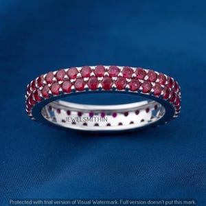 May include: A silver ring with two rows of small, round, red gemstones. The ring is set against a blue background. The text "JEWELSMITHIN" is visible on the ring.