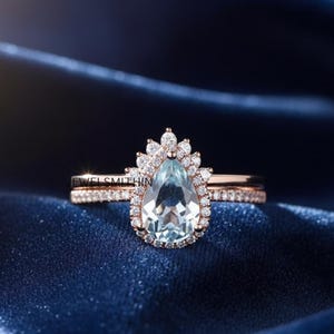 Puede incluir: Un juego de anillos de oro rosa con una piedra preciosa azul claro en forma de pera, rodeada por un halo de piedras más pequeñas. La banda también está adornada con pequeñas piedras. El juego de anillos se muestra sobre una tela azul oscuro texturizada. El texto "JEWEL SMITHIN" es visible.