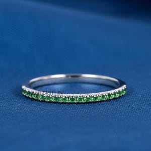 Puede incluir: Un anillo de plata con una banda de pequeñas piedras preciosas verdes redondas. El anillo está sobre un fondo azul oscuro. Las piedras preciosas están espaciadas uniformemente.