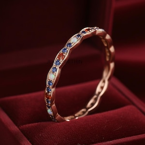 Puede incluir: Anillo de oro rosa con un diseño ondulado, engastado con piedras preciosas azules, naranjas y blancas alternas. El anillo se exhibe en un joyero de terciopelo rojo, mostrando sus intrincados detalles y colores vibrantes.