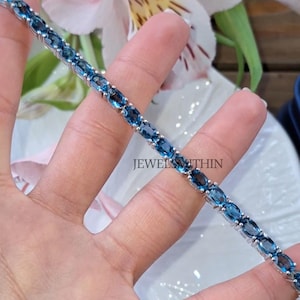 May include: A silver bracelet with oval-cut, sky-blue gemstones. The bracelet is held in a hand, with the text "JEWELS WITHIN" visible. The background includes flowers and a glimpse of a blue purse.