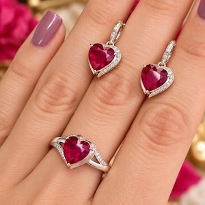 Puede incluir: Un conjunto de joyería compuesto por un anillo y dos pendientes colgantes, cada uno con una piedra roja en forma de corazón rodeada de pequeñas piedras blancas en una banda de metal plateado. El anillo tiene un diseño de banda dividida.