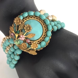 Puede incluir: Un brazalete vintage con cuentas turquesas y un medallón central dorado. El medallón tiene una gran piedra turquesa rodeada de filigrana dorada y pequeños detalles rosas y verdes. El brazalete se muestra en una mano negra.