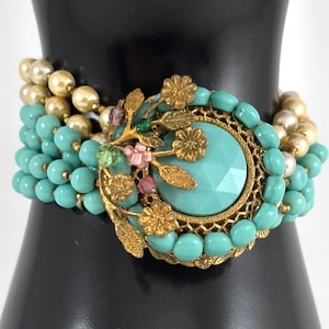 Può includere: Un bracciale a più fili con file di perline color turchese e perle color crema. Il pezzo centrale è una grande pietra turchese rotonda e sfaccettata incastonata in una montatura in metallo dorato, decorata con motivi floreali e foglie.