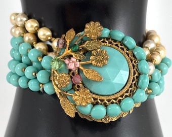 May include: A multi-strand bracelet featuring rows of turquoise-colored beads and cream-colored pearls. The centerpiece is a large, round, faceted turquoise stone set in a gold-tone metal frame, adorned with gold-tone floral and leaf accents.