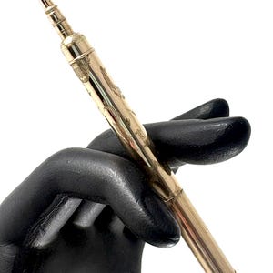 Könnte beinhalten: Ein goldfarbener Druckbleistift mit aufwendigen Gravuren. Der Stift hat ein schlankes, längliches Design mit einem kleinen Ring am Ende. Der Stift wird von einer schwarzen behandschuhten Hand vor weißem Hintergrund gehalten.