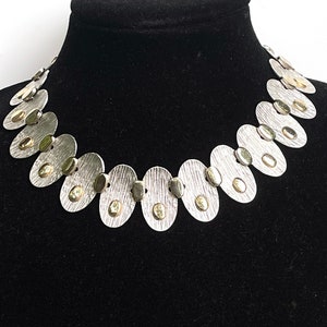 May include: A silver necklace with oval-shaped links, each with a small gold circle in the center. The necklace is on a black velvet display stand.