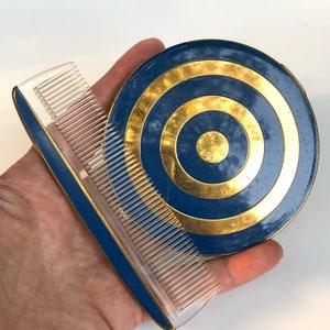 May include: A vintage vanity set comprising a blue and gold compact and a matching comb. The compact has a circular design with concentric gold rings. The comb has a clear plastic comb with a blue and gold trim. The set is likely from the mid-20th century.