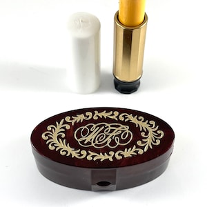 Könnte beinhalten: Ein offener Vintage-Lippenstift mit einer gelben Lippenstiftmine und einem goldfarbenen Metallgehäuse. Eine weiße Lippenstiftkappe befindet sich neben dem Lippenstift. Eine dunkelbraune ovale Kompaktpuder mit goldfarbenem floralem Filigran und den Initialen "MFK" ist ebenfalls vorhanden.
