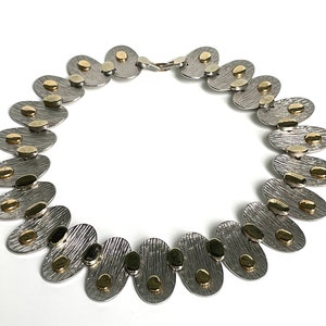 May include: A silver and gold necklace with oval-shaped links. Each link has a textured surface and is accented with small gold circles.