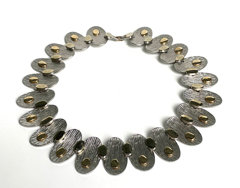 May include: A silver and gold necklace with oval-shaped links. Each link has a textured surface and is accented with small gold circles.