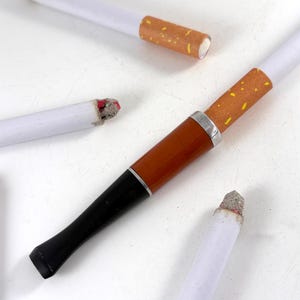 May include: An assortment of novelty cigarettes and a pipe. The cigarettes have white paper tubes with brown filters. One cigarette is attached to a brown and black pipe with a silver band. The tips of the cigarettes are gray.