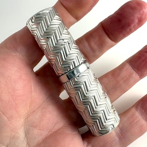 May include: A silver-colored perfume bottle with a chevron pattern. The bottle is cylindrical with a removable cap and a silver band around the middle. The bottle is held in a hand.