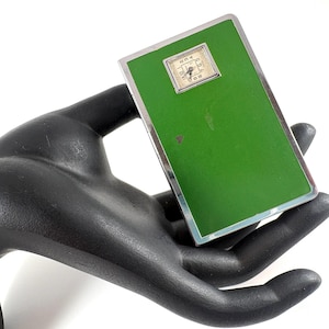May include: A rectangular, vintage-style scale with a bright green face and a small, square clock at the top. The scale has a silver-colored metal frame. The scale is held in a black hand.