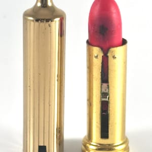 May include: A vintage lipstick tube with a gold-toned, vertically ribbed cylindrical cap and a matching gold-toned base. The lipstick itself is a vibrant, matte pink shade with a visible dark spot on the surface of the bullet.