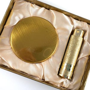 Pode incluir: Um espelho de bolso circular em tom dourado com acabamento escovado e um atomizador de perfume cilíndrico em tom dourado a condizer com superfície texturizada, apresentados numa caixa retangular forrada com tecido de cetim em tom pêssego.