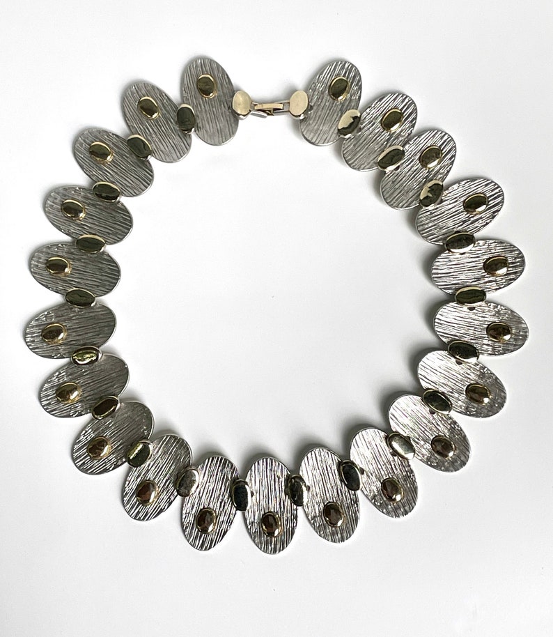 May include: A silver and gold necklace with oval-shaped links. Each link has a textured surface and is accented with small gold dots.