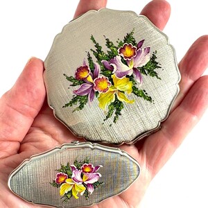 Könnte beinhalten: Zwei silberfarbene Puderdosen mit floralen Designs. Die größere Dose ist rund mit einem gewellten Rand, die kleinere oval. Beide zeigen einen handgemalten Strauß aus lila, gelben und roten Orchideen mit grünen Blättern.