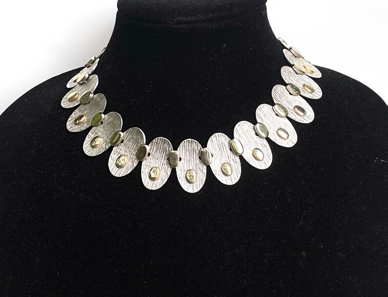 May include: A silver necklace with oval-shaped links, each with a small gold circle in the center. The necklace is on a black velvet display stand.