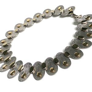 May include: A silver and gold necklace with oval-shaped links. Each link has a textured surface and is accented with small gold dots.