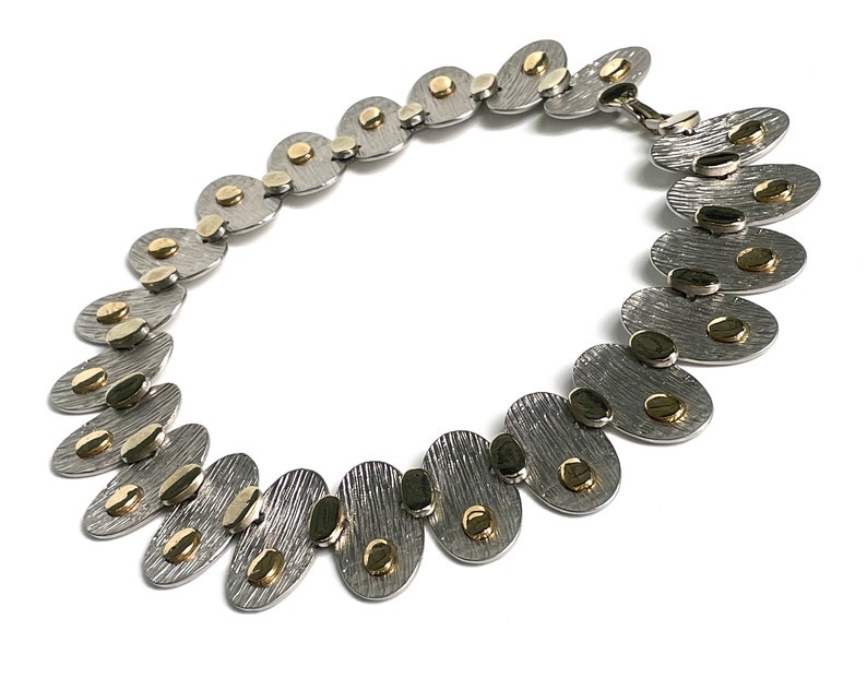 May include: A silver and gold necklace with oval-shaped links. Each link has a textured surface and is accented with small gold dots.