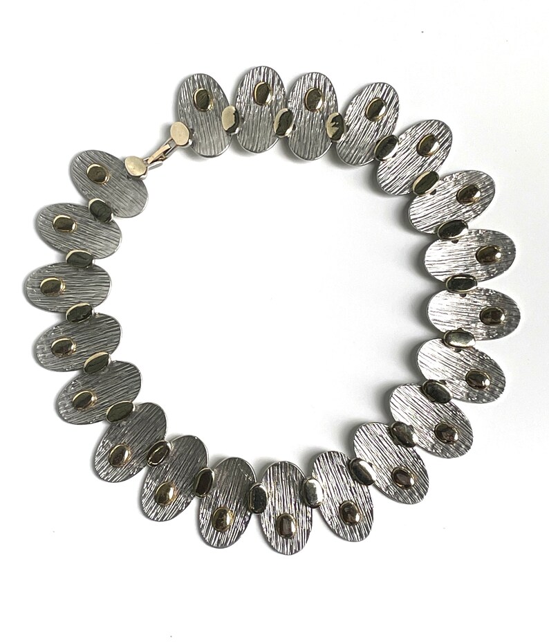 May include: A silver and gold necklace with oval-shaped links. Each link has a textured surface and gold accents.