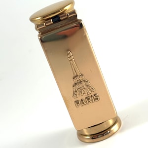 Puede incluir: Un encendedor de metal dorado con un cuerpo rectangular y extremos redondeados. La parte delantera del encendedor presenta una imagen en relieve de la Torre Eiffel y la palabra "PARIS". El encendedor está abierto, revelando el mecanismo interno.