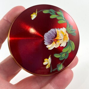 Puede incluir: Un estuche compacto redondo de color rojo metálico con un diseño floral pintado a mano. La tapa muestra un grupo de pensamientos morados y amarillos con hojas y tallos verdes, acentuado por una pequeña mariposa amarilla. El borde es dorado.