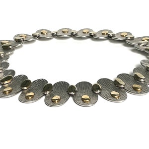 May include: A silver necklace with gold accents. The necklace is made of oval-shaped links with a textured surface. The gold accents are small, round pieces that are attached to the links.