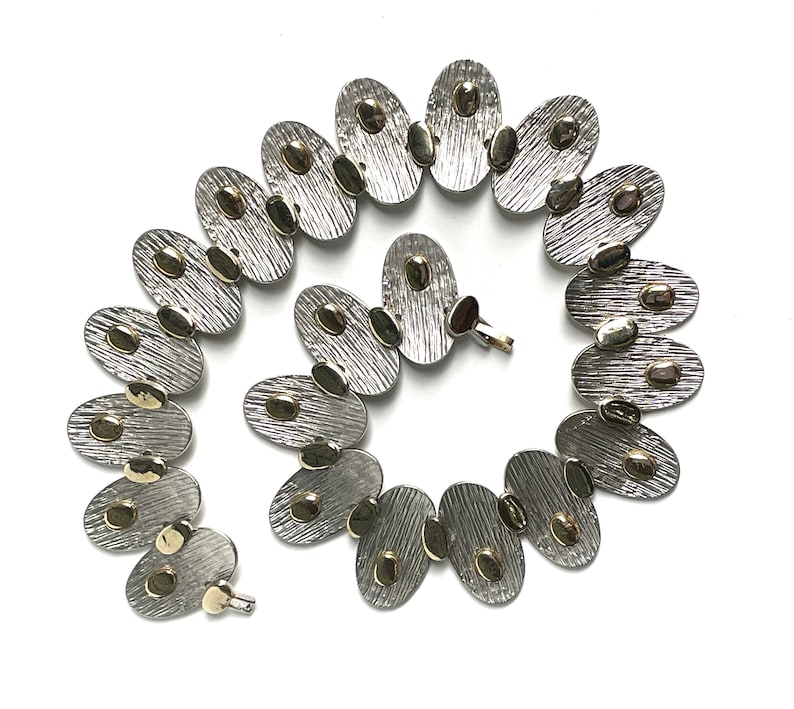 May include: A silver and gold necklace with oval-shaped links. Each link has a textured surface and is decorated with small gold dots.