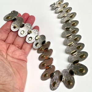 May include: A silver and gold necklace with oval-shaped links. Each link has a textured surface and is decorated with small gold dots.
