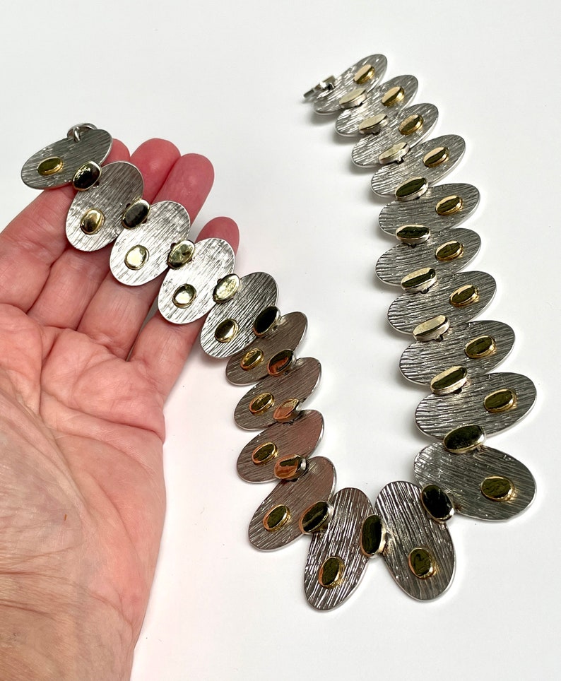 May include: A silver and gold necklace with oval-shaped links. Each link has a textured surface and is decorated with small gold dots.