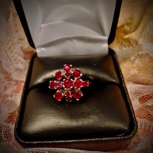 May include: A cluster ring featuring numerous round, vibrant red gemstones set in a silver-toned band. The ring is presented in a black jewellery box with a white interior, against a soft, lace-like backdrop.