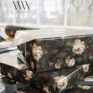Puede incluir: Dos cajas rectangulares envueltas en papel negro oscuro con un patr&oacute;n floral de rosas color crema, blancas y rojas apagadas con hojas verdes. El logotipo WW WEST WRAP es visible en la esquina superior izquierda sobre las cajas.