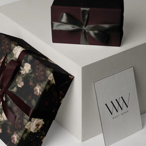 Puede incluir: Una caja de regalo rectangular envuelta en papel burdeos oscuro con una cinta de sat&eacute;n gris oscuro, una caja m&aacute;s grande con estampado floral oscuro y cinta burdeos, y una tarjeta blanca con el texto negro WW WEST WRAP.