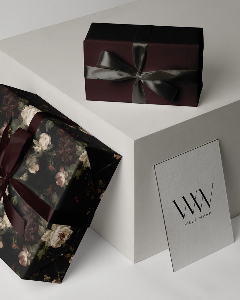 Puede incluir: Una caja de regalo rectangular envuelta en papel burdeos oscuro con una cinta de sat&eacute;n gris oscuro, una caja m&aacute;s grande con estampado floral oscuro y cinta burdeos, y una tarjeta blanca con el texto negro WW WEST WRAP.