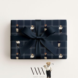 May include: A navy blue gift box wrapped in patterned paper featuring a repeating design of teddy bears in golf attire. The box is tied with a matching navy blue satin ribbon and bow. The West Wrap logo is at the bottom.