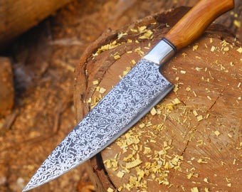 May include: A handcrafted chef's knife with a wooden handle and a patterned blade. The blade has a silver finish with a decorative swirling design. The handle is a light brown color. The knife is resting on a wooden surface.