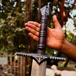 Könnte beinhalten: Ein dekoratives Schwert mit einem dunkelbraunen Griff und einer silbernen Klinge. Der Griff hat oben ein geometrisches Design und wird von einer Hand gehalten. Der Parierstange und die Klingenbasis des Schwertes weisen aufwendige silberne Details auf.