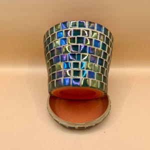 May include: A mosaic-tiled candle holder with a cylindrical shape, featuring iridescent blue, green, and purple tiles. The holder sits atop a matching saucer, both with a warm orange interior.