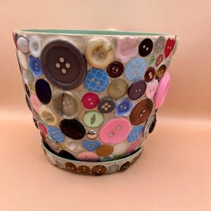 May include: A flower pot decorated with a mosaic of colourful buttons. The buttons are in shades of brown, pink, blue, and white, creating a textured surface. The pot has a matching saucer and a light green interior.