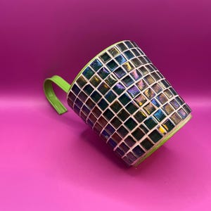 May include: A mosaic-tiled mug with a green handle and rim. The mug's body is covered in small, iridescent, square tiles that reflect multiple colours. The handle and rim are a bright green colour. The mug is set against a pink background.